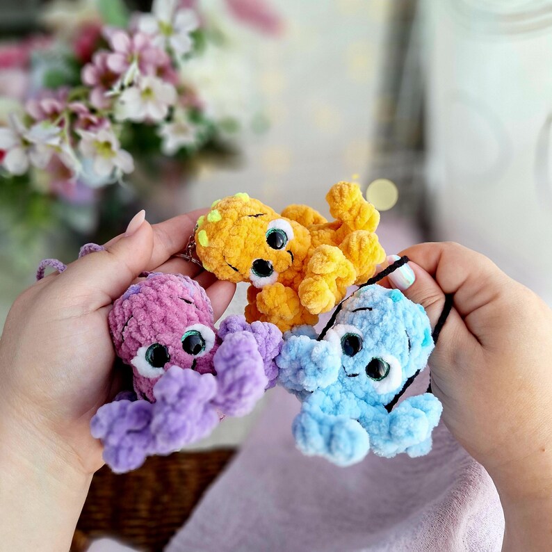 May include: Three crocheted octopus keyrings in purple, yellow, and blue. Each has large, googly eyes and a black string loop. The octopus toys are made from soft, plush material, ideal for attaching to keys or bags.