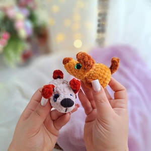 May include: Two crocheted plush toys in the shape of dogs. One dog is light pink with red ears and a black nose. The other dog is orange with brown ears and a green eye.