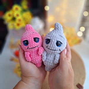 May include: Two crocheted ghost-shaped plush toys. One is pink with a toothy grin and the other is gray with a more subtle smile. Both have large blue eyes.