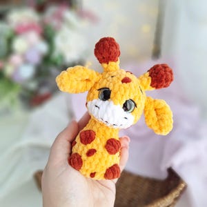 May include: A crocheted giraffe toy in yellow and brown, featuring red spots. The giraffe has large eyes, a white muzzle, and small ears. The soft toy is held in a hand, highlighting its texture and detailed design. A charming handmade item.
