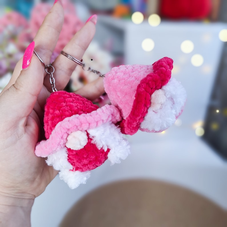 May include: Two pink and white crocheted gnome keychains with white beards and pink hats. The keychains are attached to a silver key ring.