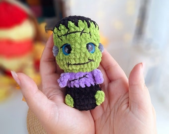 May include: A green and purple crocheted Frankenstein monster toy with big blue eyes. The toy is being held in a person's hands.