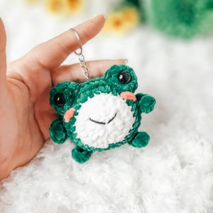 May include: A green crocheted frog keychain with white yarn for the face and black eyes. The frog has a small pink mouth and is holding a silver key ring.