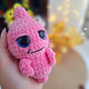 May include: A pink crocheted amigurumi toy with large blue eyes and a small smile. The toy is made of soft yarn and has a fuzzy texture.