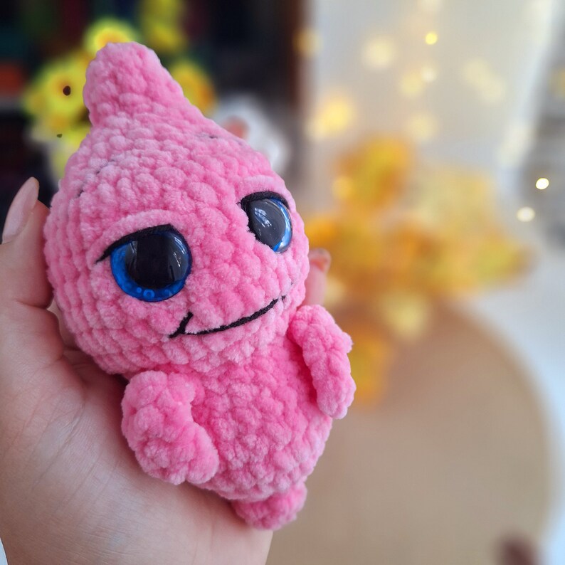 May include: A pink crocheted amigurumi toy with large blue eyes and a small smile. The toy is made of soft yarn and has a fuzzy texture.