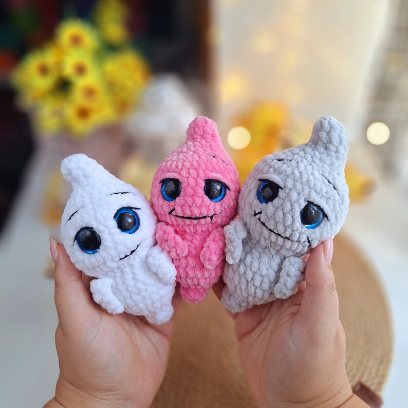 May include: Three crocheted ghost-shaped plush toys with embroidered eyes. The ghosts are pink, white, and gray.