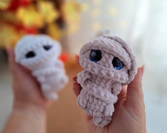 May include: Two crocheted plush toys in the shape of mummies. The mummy on the left is white and the mummy on the right is light purple. Both mummies have large blue eyes.