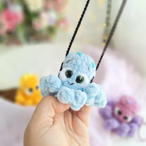 May include: A light blue crocheted octopus pendant with large, expressive eyes and a smiling face. The plush octopus is suspended from a black cord. Other crocheted octopus toys in yellow and purple are visible in the background.