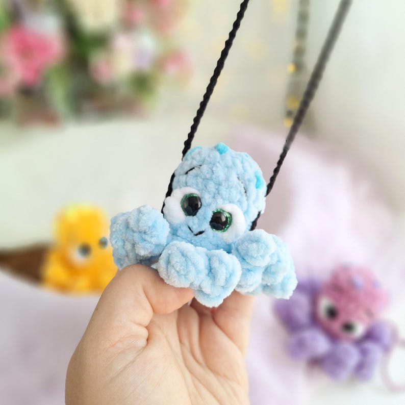 May include: A light blue crocheted octopus pendant with large, expressive eyes and a smiling face. The plush octopus is suspended from a black cord. Other crocheted octopus toys in yellow and purple are visible in the background.