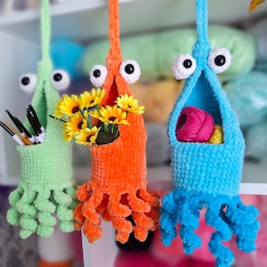 May include: Three crocheted hanging baskets with whimsical octopus-like legs. The baskets are in green, orange, and blue. One basket holds a bouquet of sunflowers, another holds balls of yarn, and the third is empty.