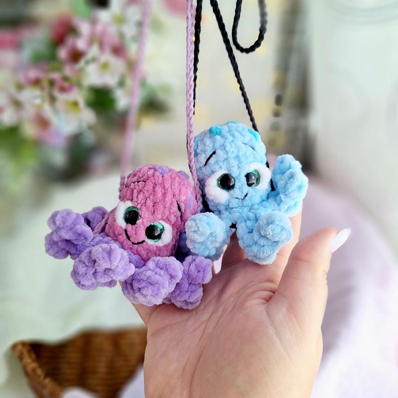 May include: Two crocheted octopus toys, one in pink and purple, the other in blue. Each has large eyes and a smiling face. They hang from black and pink cords, possibly for wearing or display. The toys are soft and fluffy.