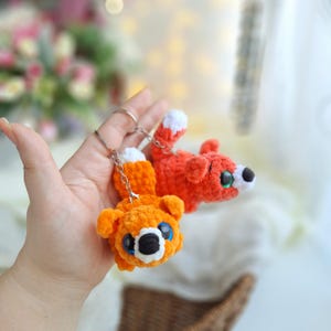 Op de afbeelding: Twee gebreide vos sleutelhangers, een oranje en een rode, met witte accenten op de staarten. De sleutelhangers worden in een hand gehouden.