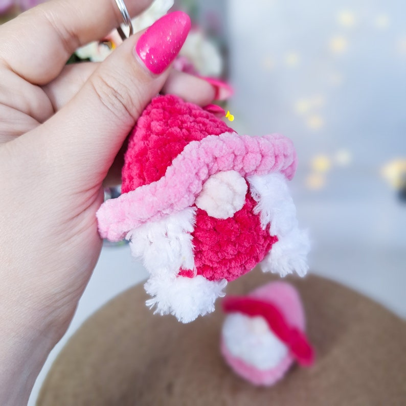 May include: A pink and white yarn gnome keyring with a white beard. The gnome is holding a small white heart.
