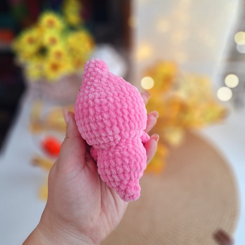 May include: A pink crocheted plush toy in the shape of a small animal. The toy is soft and fuzzy and has a textured surface.