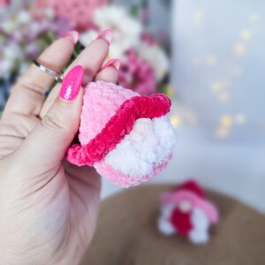 May include: A pink and white crocheted gnome keyring with a red hat. The gnome is being held in a hand with pink nail polish.