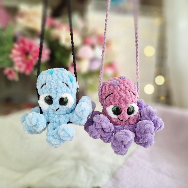 May include: Two crocheted octopus toys, one in light blue and the other in purple, with large googly eyes and smiling faces. Each has a matching strap for hanging. The toys are made of soft, textured yarn.