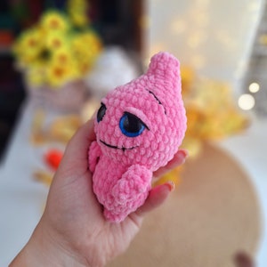 May include: A pink crocheted plush toy with blue eyes. The toy is shaped like a teardrop and has a small smile.