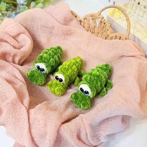 Puede incluir: Tres juguetes de cocodrilo de ganchillo verde hechos a mano con ojos blancos y azules descansan sobre una tela rosa. Los caimanes tienen un diseño texturizado y con bultos y están dispuestos en fila. El fondo incluye una cesta tejida y detalles florales.