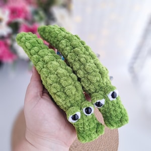 May include: Two green crocheted alligator toys with black eyes and white pupils. The alligators are lying on a brown surface.