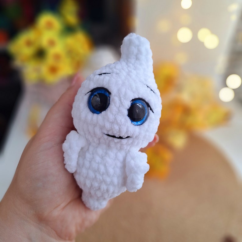 May include: A white crocheted ghost plush toy with blue eyes. The ghost has a small smile and is holding its arms out.
