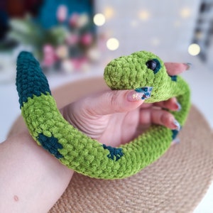 May include: A green and blue crocheted snake toy. The snake is curled up in a person's hand.