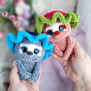 May include: Two handmade crochet dolls. One is grey with a blue ruffled headpiece, the other is pink with a green and red ruffled headpiece. Both dolls have smiling faces, blue eyes, and are held in hands.