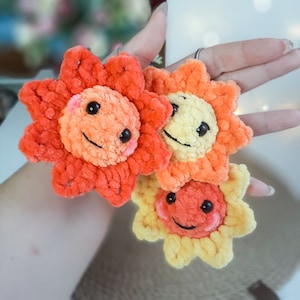 Puede incluir: Tres adornos de crochet con forma de sol con caras sonrientes. El sol más grande es naranja y rojo, el sol del medio es amarillo y naranja, y el sol más pequeño es amarillo.