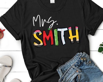 May include: Black t-shirt with the text "Mrs. SMITH" in colorful letters. The letters are decorated with images of pencils, rulers, and an apple.