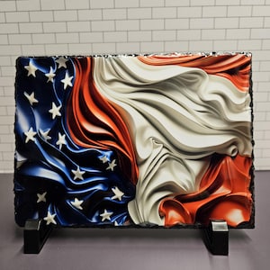 Puede incluir: Placa rectangular de pizarra con un diseño estilizado de la bandera estadounidense. El diseño presenta estrellas y rayas en azul, blanco y rojo, con un efecto texturizado y fluido. La placa está soportada por dos soportes negros.