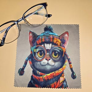 May include: A pair of black-framed eyeglasses resting on a square microfiber cleaning cloth. The cloth features a colorful illustration of a cat wearing a patterned winter hat, a matching scarf, and bright orange-rimmed glasses.