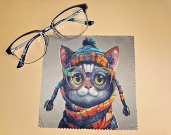 Puede incluir: Un par de gafas con montura negra sobre un paño de limpieza de microfibra cuadrado. El paño presenta una ilustración colorida de un gato con un gorro de invierno estampado, una bufanda a juego y gafas con montura naranja.