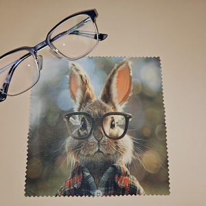 Puede incluir: Un par de gafas con montura azul oscuro descansa sobre un paño de microfibra. El paño presenta un conejo con gafas y una camisa a cuadros. El fondo del paño es una escena borrosa al aire libre.