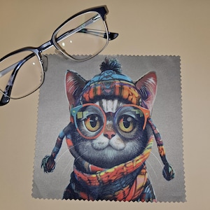 May include: Black and silver eyeglasses are positioned above a grey microfiber cleaning cloth. The cloth displays a colourful illustration of a cat wearing glasses, a hat, and a scarf. The cat's glasses are orange and blue.