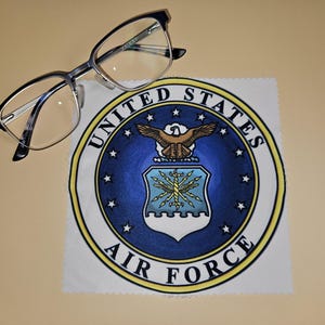 May include: A pair of eyeglasses rests on a cleaning cloth displaying the United States Air Force emblem. The emblem is a circular design with a blue background, a central crest, and the words "UNITED STATES AIR FORCE".