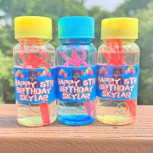 May include: Three bubble bottles with yellow, blue, and turquoise lids. Each bottle has a red bubble wand and a red plastic toy inside. The bottles have a blue label with the text "HAPPY 5TH BIRTHDAY SKYLAR" and colourful balloons.