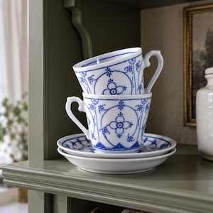 May include: Two white porcelain teacups with blue floral patterns stacked on top of each other, resting on a matching white saucer. The cups feature delicate blue line work and circular motifs, with a curved handle on the side of each cup.