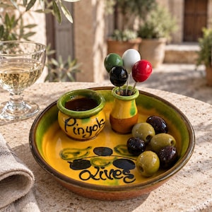 May include: A round yellow and green ceramic serving dish with the text Pinyols Olives painted in black. The dish includes a small attached bowl, a toothpick holder with four colored picks, and several scattered green and black olives.