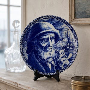 Può includere: Un piatto decorativo rotondo in ceramica con un'illustrazione blu e bianca di una persona barbuta che indossa un cappello e fuma la pipa. Lo sfondo raffigura una scena costiera con una barca a vela. Il piatto presenta un bordo decorato.