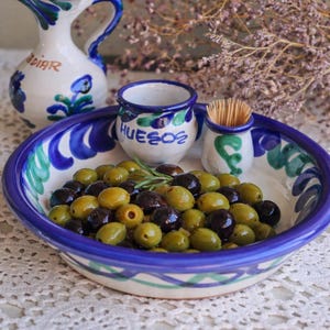 May include: A ceramic bowl filled with green and black olives, a small jug, and two small ceramic containers. The bowl and jug have blue and green painted designs. The container has the word "HUESOS" on it.