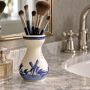 Könnte beinhalten: Eine cremefarbene Keramikvase mit einem handgemalten blauen Windmühlen- und Landschaftsmotiv, die fünf Make-up-Pinsel mit dunkelblauen Griffen und braunen Borsten enthält. Die Vase hat einen schmalen Hals und einen ausgestellten Rand.