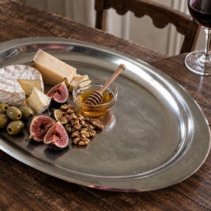 Peut inclure: Un plateau de service ovale en métal argenté contenant une sélection d'aliments, notamment une meule de brie, des morceaux de fromage à pâte dure, des olives vertes, des figues fraîches coupées, un bol de miel avec une cuillère en bois et des noix.