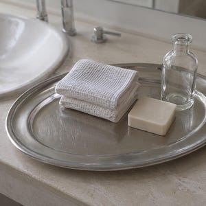 Peut inclure: Un plateau ovale en métal argenté contenant une pile de deux gants de toilette blancs pliés à texture gaufrée, un pain de savon rectangulaire blanc cassé et une petite bouteille en verre transparent vide au goulot étroit.