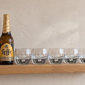 May include: A brown glass beer bottle with a gold foil neck label and a yellow label featuring the text Leffe Blonde Blond, positioned next to five clear glass tumblers, each etched with the Leffe logo and featuring a thick, bubble-textured base.