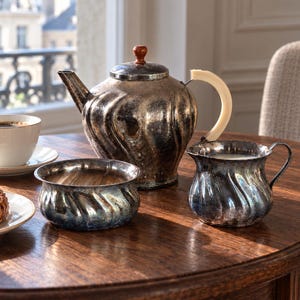 Op de afbeelding: Een verzilverd theeservies bestaande uit een theepot met een lichtgekleurd handvat en een bruine knop, een suikerpot en een melkkannetje, alle met een golvend ribbelpatroon en zichtbare aanslag, inclusief een witte keramische kop en gebak.