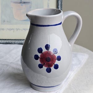 Può includere: Brocca in ceramica bianca con una striscia blu e un motivo floreale rosso e blu. La brocca ha un manico e un beccuccio. Sullo sfondo, un'immagine incorniciata di un albero in vaso.
