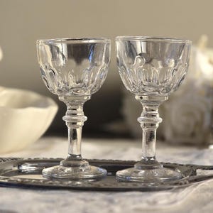 May include: Two clear, ornate crystal wine glasses sit on a silver tray. The glasses have detailed patterns and are displayed with a white cherub statue and a silver sugar bowl, suggesting a vintage or antique theme.
