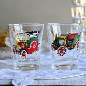 Puede incluir: Dos vasos de chupito transparentes con diseños de coches antiguos coloridos. Uno es rojo, verde y amarillo, el otro verde, rojo y amarillo. Los vasos están sobre un paño blanco, con un plato de cristal decorativo y una jarra de cristal transparente al fondo.