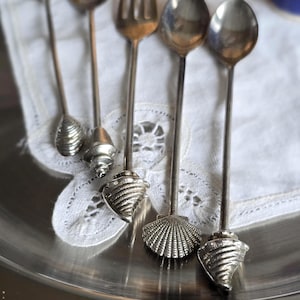 May include: A collection of silver-toned serving utensils with seashell-shaped handles. The set includes spoons and a fork, arranged on a silver tray and a white lace doily. The utensils are likely for serving appetizers or desserts.