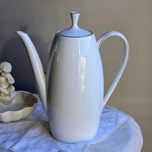 Op de afbeelding: Een witte porseleinen koffiepot met een gebogen handvat en een lange tuit. De pot heeft een deksel met een klein handvat en een dunne zilveren rand. Hij staat op een witte doek met een kanten rand. Een klein cherubijnbeeldje is zichtbaar op de achtergrond.