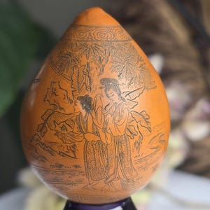 May include: An orange gourd-shaped ornament with a detailed black etched design. The design shows two figures in traditional clothing in a landscape of trees and foliage. The gourd is mounted on a dark purple base.